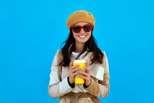 Emotional happy beautiful woman in beige coat beret and sunglasses on blue background drinking coffee in paper cup smiling, morning food, joyful drinks emotions people beauty fashion lifestyle concept