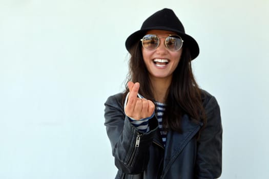 Happy stylish female in hat and leather jacket showing heart gesture with fingers while looking at camera on light blue background