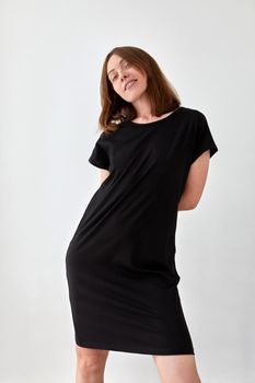 Delighted young slim female in casual black dress standing on white background in studio and looking at camera