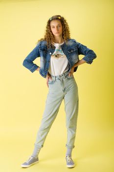 Full length shot of girl with hands on her waist. Curly blonde girl in trendy denim outfit posing against light yellow background. Teenager wearing jeans, denim jacket and white t-shirt