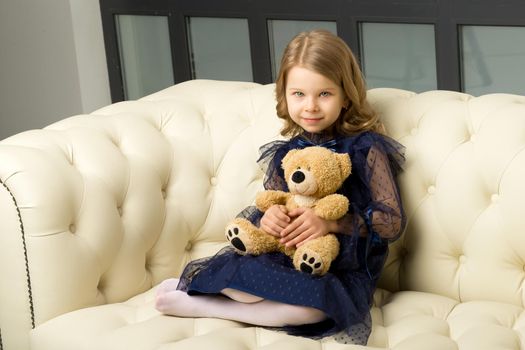 Lovely little girl hugging her teddy bear toy. Adorable blonde girl wearing trendy clothes sitting on white leather comfortable sofa and smiling at camera.