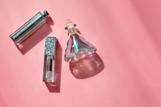 Top view of various shiny bottles with perfume and lipsticks arranged on pink table lit by sunlight