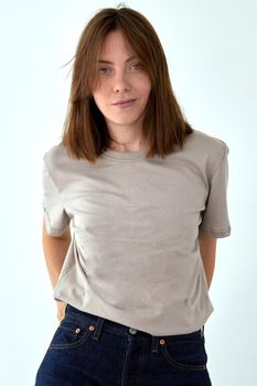 Positive female model wearing gray t shirt and jeans standing with hands behind back against white background and looking at camera