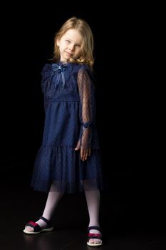 Cute charming little girl in elegant blue dress. Full length portrait of adorable blonde girl wearing nice blue lace dress posing on black background in studio.