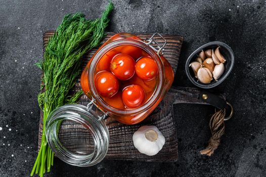 Pickled cherry tomatoes in a glass jar with herbs. Black background. Top view.