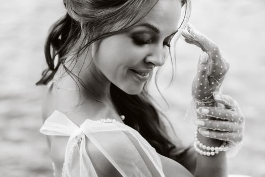 An elegant bride in a white dress and gloves is sitting by the lake in the Park, enjoying nature.A model in a wedding dress and gloves in a nature Park.Belarus.black and white photo.