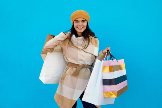 Cute attractive happy smiling stylish woman shopaholic in trendy beige plaid coat and beret holding shopping bags on blue studio background isolated, colorful sale excited fashion trend outdoors