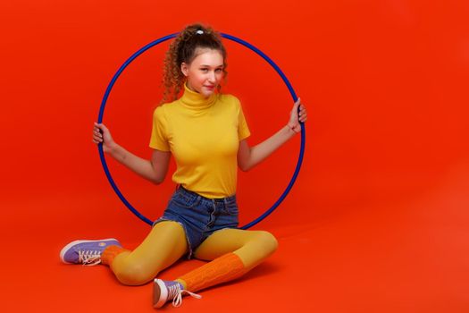 Shot of pretty fitness girl holding hula hoop. Portrait of smiling girl doing exercises with blue hoop. Student looking stylish in turtleneck blouse, leggins, shorts and socks gaiters
