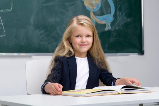 Happy schoolgirl sitting at desk. Elementary school student in blue jacket and white blouse reading book on background of blackboard in classroom. Back to school, education concept