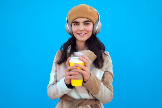 Cheerful positive millennial woman with headphones wear beret and beige coat looking at camera, holding paper cup of hot coffee or tea takeaway outside pastel blue background