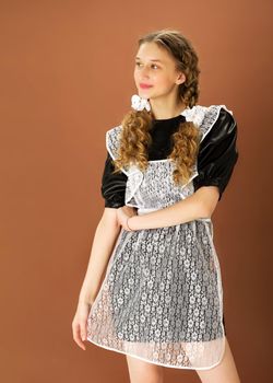 Cheerful blonde teen girl in retro school uniform. Portrait of graduate girl with two curly braids wearing USSR style school uniform posing against light brown background.