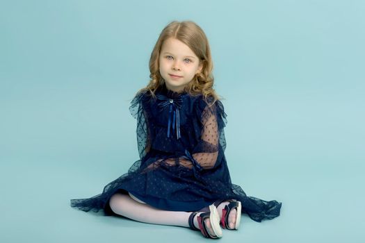 Happy little girl in a blue dress sitting on the floor. Portrait of a charming blonde in a beautiful lace dress sitting on a light blue background in the studio.