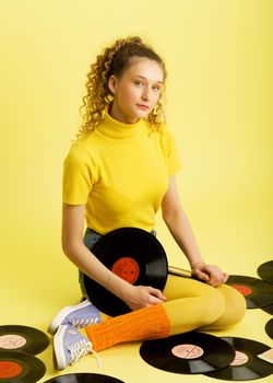 A beautiful girl is sitting on the floor with vinyl records. Portrait of happy curly haired girl in bright yellow turtleneck and leggings listening to music and relaxing on isolated yellow background