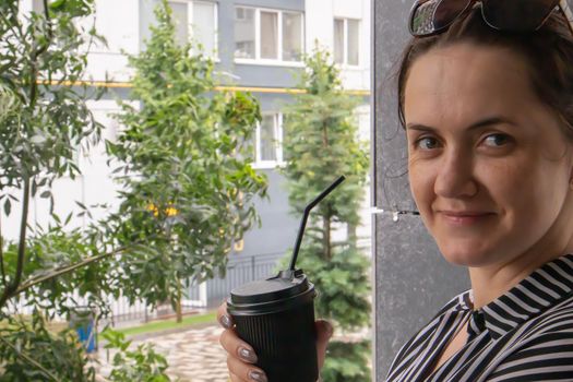 One black paper cup with a lid and a straw in the hands of a young happy smiling woman outdoors in summer in sunny weather on the background of houses, in a coffee shop or restaurant. Coffee to go