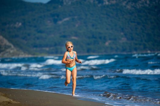 A little girl in a blue swimsuit and glasses runs on a Mediterranean beach in Turkey.