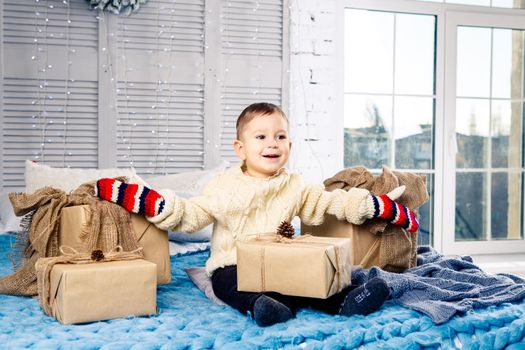 little funny playful boy a child sits on a bed on Christmas day with gift boxes in white wool knitted sweater and big bright mittens on it and laughs out loud. In interior there is a festive decor.