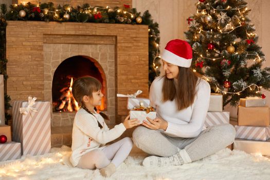 Young woman gets Christmas gift from her cute little daughter with two pigtails, at Christmas morning in living room. Celebrating Christmas and New Year, having fun, happy time together.