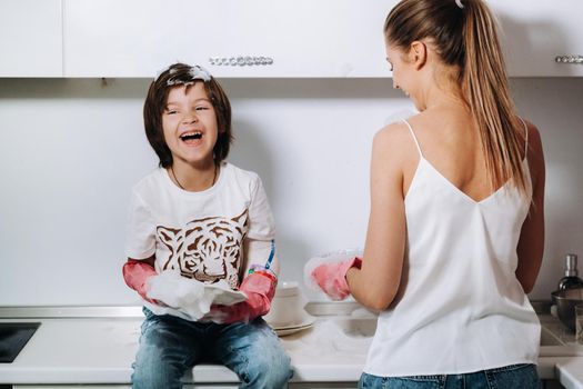 housewife mom in pink gloves washes dishes with her son by hand in the sink with detergent. A girl in white and a child with a cast cleans the house and washes dishes in homemade pink gloves.A child with a cast washes dishes and smiles.