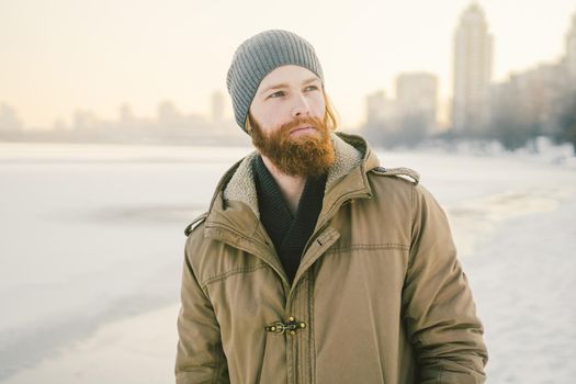 Close-up Caucasian young male red hair and beard in a hat and a park coat posing winter model against a background of a lake to freeze snow.