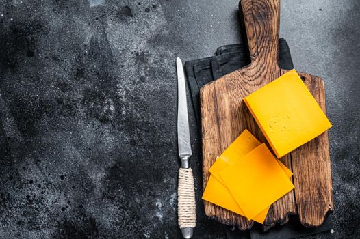 Slices of Cheddar Cheese on a wooden cutting board. black background. Top view. Copy space.