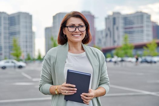 Outdoor portrait of business woman 40s of age. Smiling female in glasses with business notebook in hands, city buildings urban architecture background
