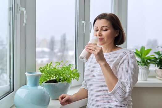 Middle-aged attractive woman drinking dairy product, yogurt, milk in bottle. Female at home near window, winter snowy day, copy space. Diet, healthy food, nutrition concept