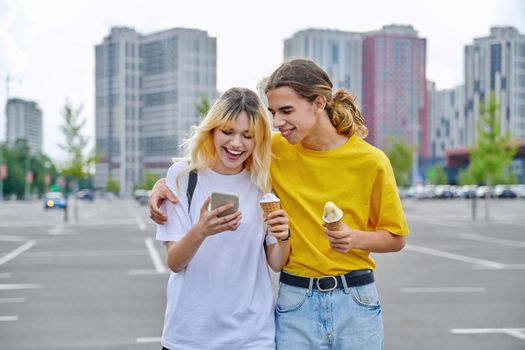 Happy laughing couple of teenagers walking hugging together in city, guy and teenage girl with ice cream looking into smartphone, friendship, relationship, summer, leisure, lifestyle, adolescence