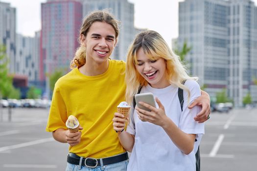 Happy laughing couple of teenagers walking hugging together in city, guy and teenage girl with ice cream looking into smartphone, friendship, relationship, summer, leisure, lifestyle, adolescence