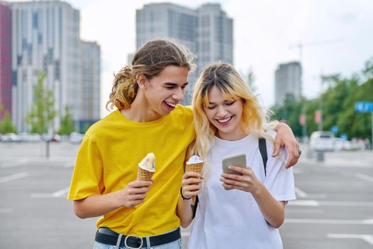 Happy laughing couple of teenagers walking hugging together in city, guy and teenage girl with ice cream looking into smartphone, friendship, relationship, summer, leisure, lifestyle, adolescence