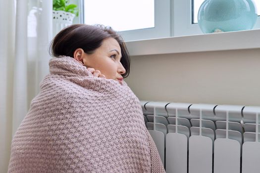 Winter, heating season. A frozen woman in warm woolen sweater under knitted blanket sitting in home room near heating radiator