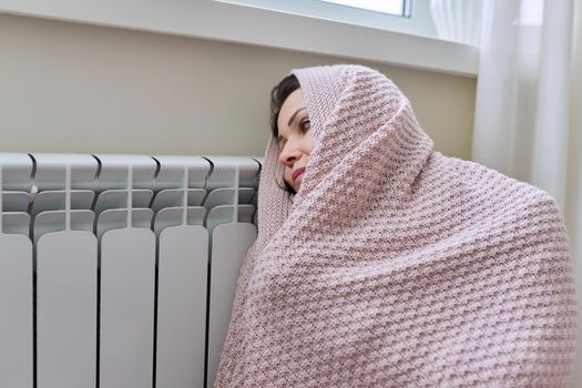 Winter, heating season. A frozen woman in warm woolen sweater under knitted blanket sitting in home room near heating radiator