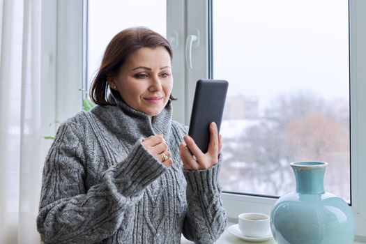 Middle-aged woman in a warm knitted sweater with a smartphone in her hands, home window with winter snowy landscape background, copy space