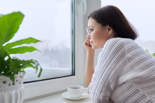Season winter, snowy day, middle aged smiling woman with cup of coffee looking out the window. Female 40s of age, lifestyle, portrait, copy space