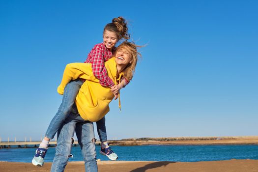 Happy mom and daughter child on seashore, relaxing on sandy beach, autumn winter spring season, copy space, blue sky background. Family vacation, travel, parent child relationship, happiness, joy