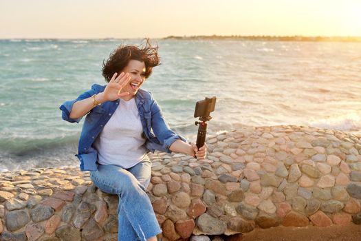 Technology, lifestyle, leisure and travel. Middle-aged female making online video call talking laughing, using smartphone, sunset seascape, sandy beach background, tropical autumn winter spring season