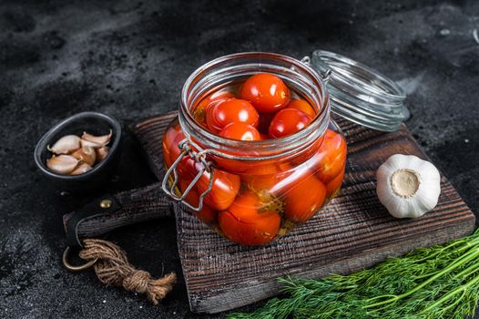 Pickled cherry tomatoes in a glass jar with herbs. Black background. Top view.