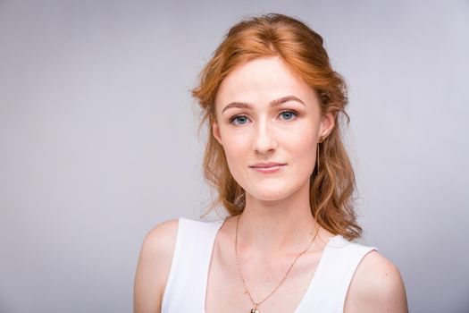 Closeup portrait young, beautiful business woman, student with lred, curly hair and freckles on face on gray background in the studio. Dressed in white blouse with short sleeves about open shoulders.