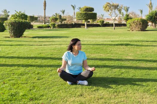 Middle aged woman sitting on grass in lotus position and meditating, sunny morning on summer day. Meditation, relaxation, healthy lifestyle and body, mental health, people 40s