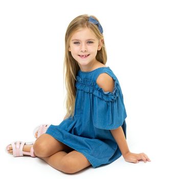 Charming little girl sitting on floor. Smiling blonde girl of six or seven years old wearing blue denim dress and sandals sitting on her knees on isolated white background and looking at camera
