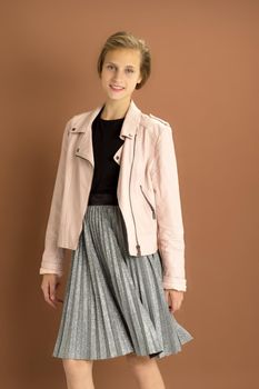 Happy girl whirling against brown background. Joyful attractive brown haired teen girl dressed pink leather jacket and gray fluttering pleated skirt having fun in studio