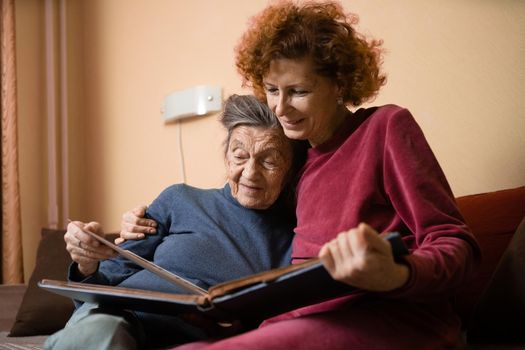 Senior woman and her adult daughter looking at photo album together on couch in living room, talking joyful discussing memories. Weekend with parents, family day, thanksgiving, mom's holiday.