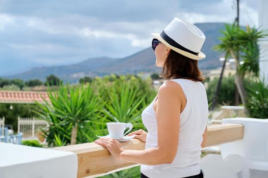 Tourism, travel, vacation, resort. Happy beautiful middle age woman in sunglasses hat with cup of coffee enjoying beautiful view of mountain sky mediterranean natural landscape, copy space