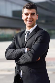 Young businessman near a modern office building wearing black suit and tie. Man with blue eyes smiling.
