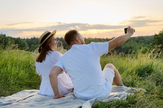 Beautiful adult couple together taking selfie photo on smartphone. Happy man and woman sitting on grass outdoors in sunset sunlight. Relationships, love, date, people 30s 40s age