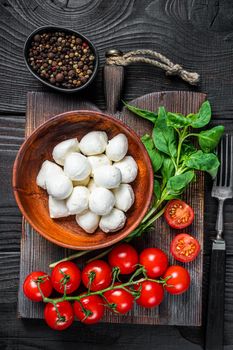 Italian mini Mozzarella cheese balls, basil and tomato cherry ready for cooking Caprese salad. Black wooden background. Top view.