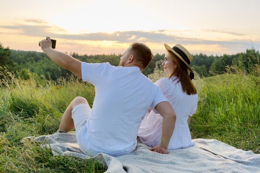 Beautiful adult couple together taking selfie photo on smartphone. Happy man and woman sitting on grass outdoors in sunset sunlight. Relationships, love, dating, people 30s 40s age