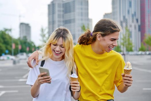 Happy laughing couple of teenagers walking hugging together in city, guy and teenage girl with ice cream looking into smartphone, friendship, relationship, summer, leisure, lifestyle, adolescence