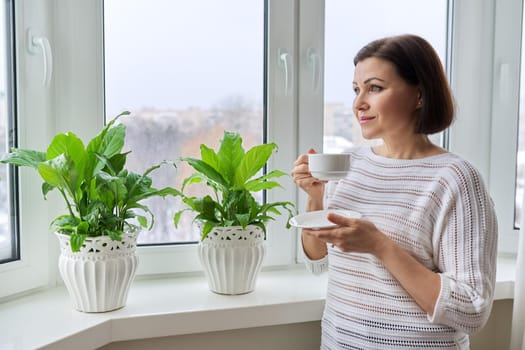 Season winter, snowy day, middle aged smiling woman with cup of coffee looking out the window. Female 40s of age, lifestyle, portrait, copy space