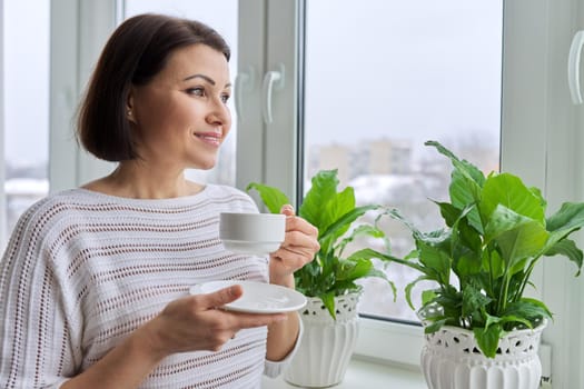 Season winter, snowy day, middle aged smiling woman with cup of coffee looking out the window. Female 40s of age, lifestyle, portrait, copy space