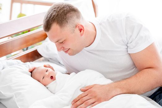 Young father sitting in the bed and looking with tenderness at his newborn baby daughter. Smiling parent dad and infant child girl at home with daylight